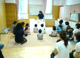 子どもたちと大人たちが床に座っていて、担当の職員がボードを見せながら何か説明をしている写真