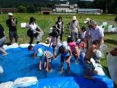 子供たちがプールに入り、周りで保護者たちが様子を見ている、魚道見学イベントでのニジマスのつかみ取りの写真