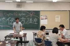 室内で3人が座り、1人が文字が書かれた黒板を背に立っている写真