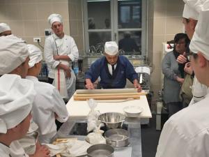 調理場で真剣な表情でそばを打つ男性とその様子を周りで興味深そうに見学する沢山の人々の写真