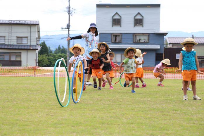 青いエプロンを付けた女性の前で遊んでいる麦わら帽子をかぶった幼児たちの写真