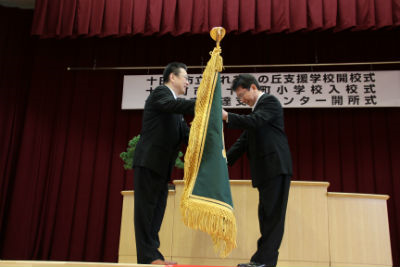 2人の大人が壇上で旗を受け渡している。