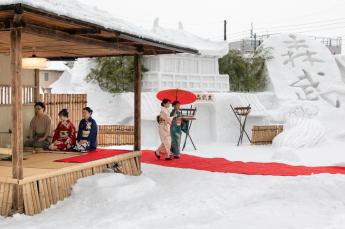 雪まつりの中赤い歩行路を赤い傘を指して進む着物の女性2人と、左側で野点をしている和服の3人組の様子の写真