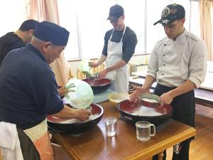 日差しが入る明るい部屋で、男性4人が大きな器に粉を入れ蕎麦を作っている写真