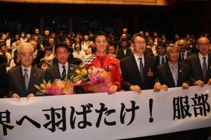 凱旋報告会にて応援用横断幕を手に記念撮影に応じる、服部勇馬選手と市長および役員たちの写真