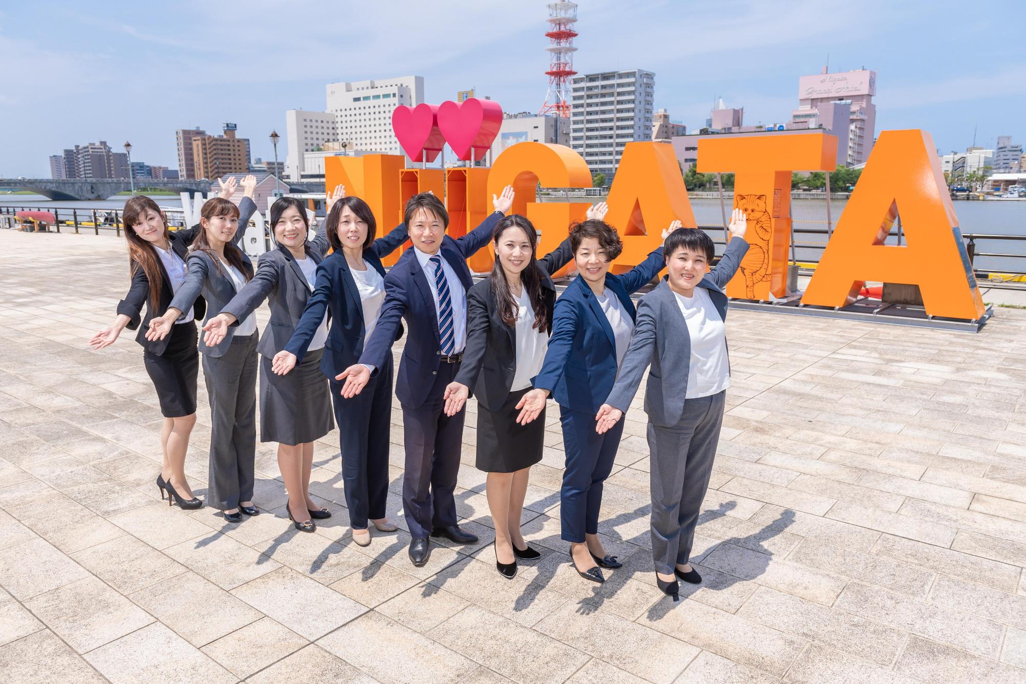 河川の向かい側にはビル群が見え、川沿いにオレンジの字体で「新潟」の文字が英語になったモニュメントの前で、並んで両手を斜めに広げているスーツ姿の男女の写真