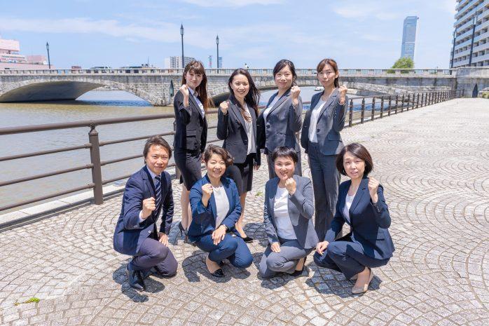 奥に橋が見え、川沿いの石畳の道でガッツポーズをして並ぶ、スーツ姿の男女の写真