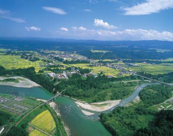 青々とした水田地帯に、大きな川と支流が合流した地点に広がる市街地の写真