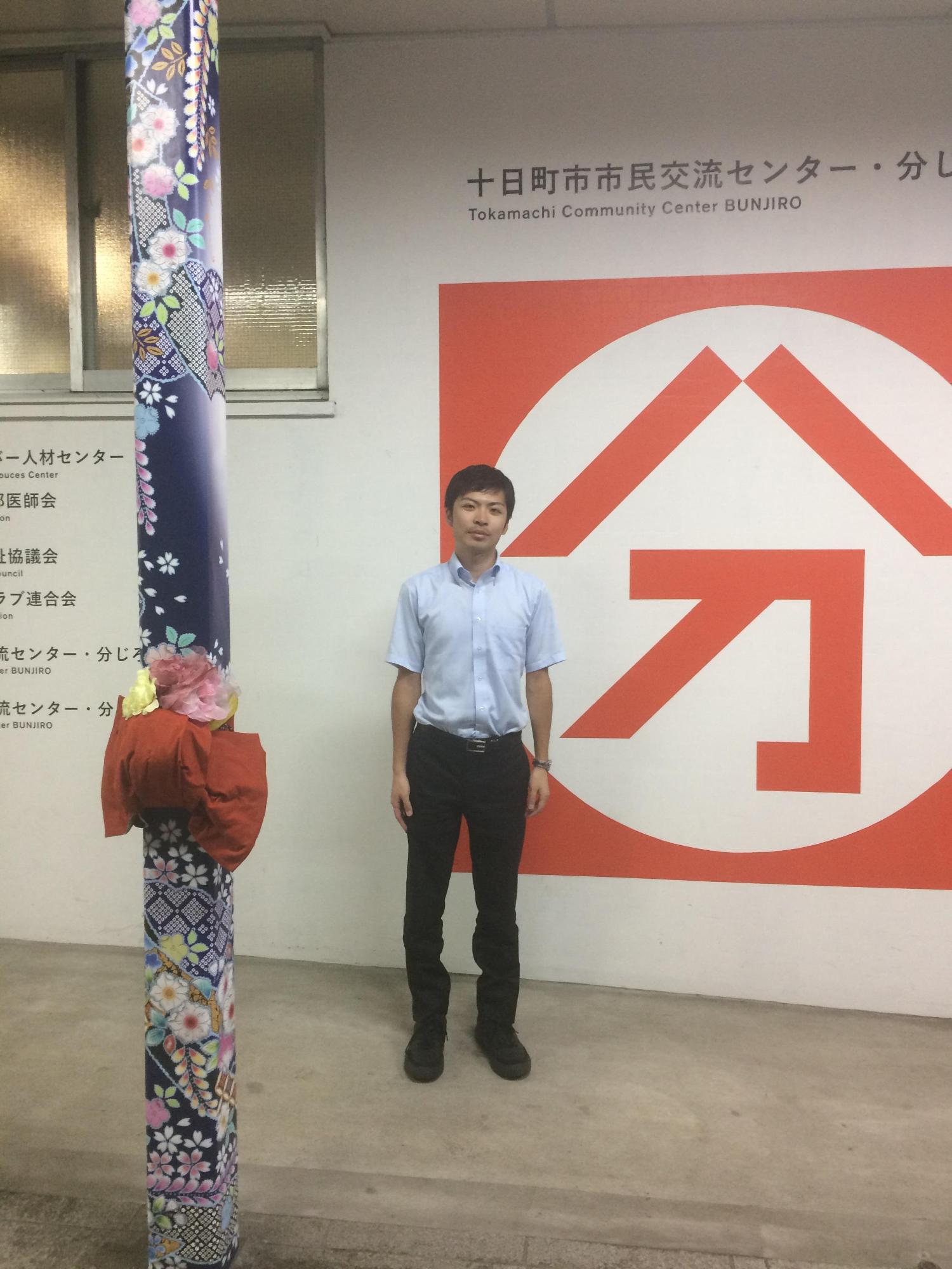 十日町市市民交流センターの赤い文字アートの入った壁際に立つ若い男性と、紫の着物の生地が巻かれて花や赤い帯があしらわれた柱が立っている写真