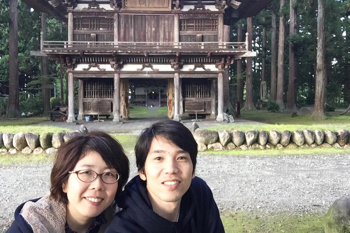古い木造で出来た大きな仁王門の前で、黒いパーカーを着た男性と眼鏡姿の女性が笑顔で正面を向いて記念撮影をしている写真