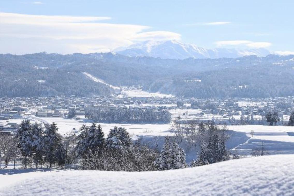遠くには山々が見え、ふもとには一面に雪が降り積もった町や木々がある豊かな風景を高台から撮影した写真