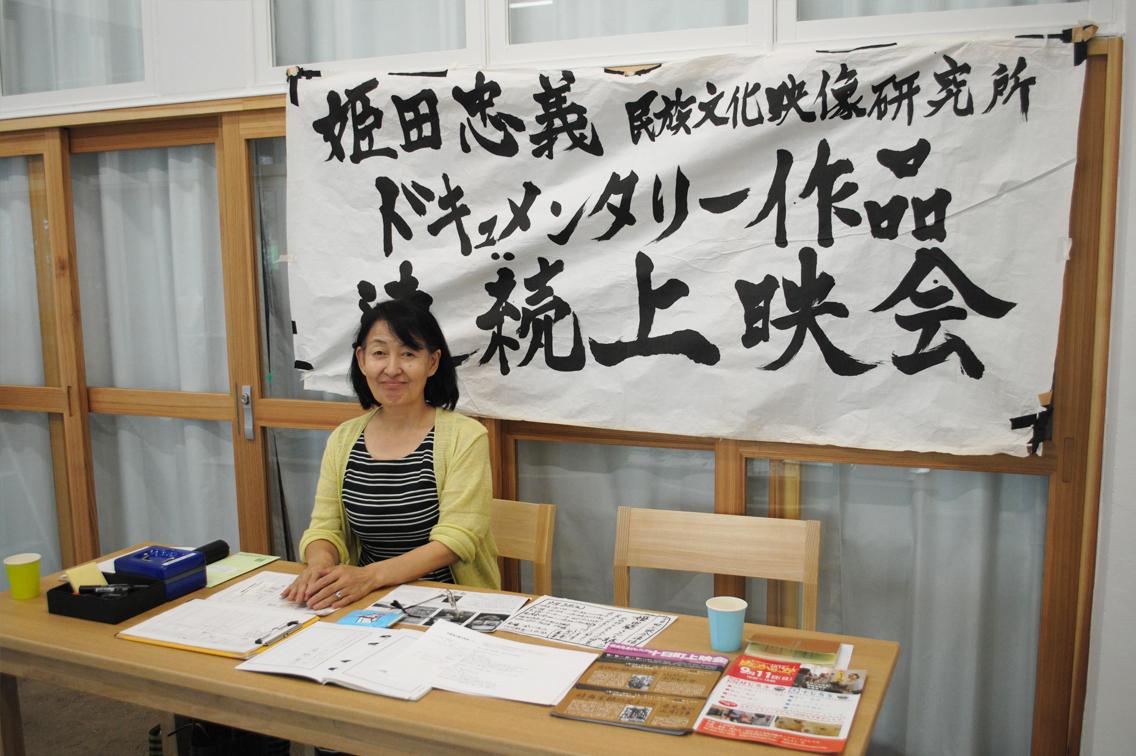 迫力ある墨字で書かれた上映会の布の旗の前で、資料が並べられた木のテーブルに座り、こちらに向かって穏やかにほほ笑む中年女性の写真
