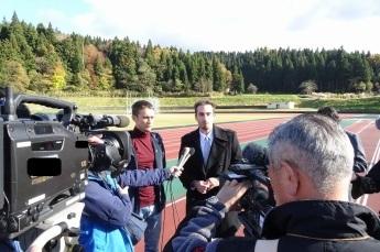 陸上競技場で撮影機材を向けられながらインタビューに答える男性の写真