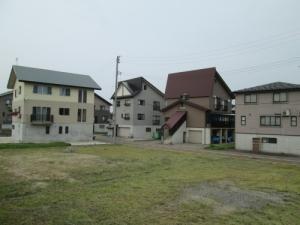 うっすら草が生えている空き地の奥に、一戸建ての住宅が立ち並んでいる写真