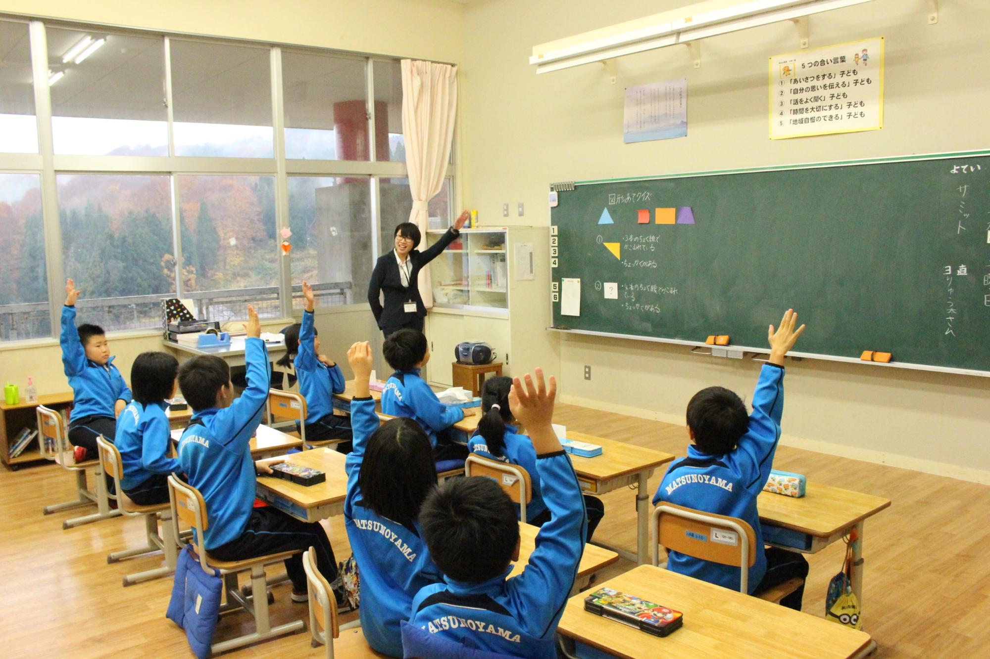 教室で黒板に向かい挙手をしている大勢の小学生