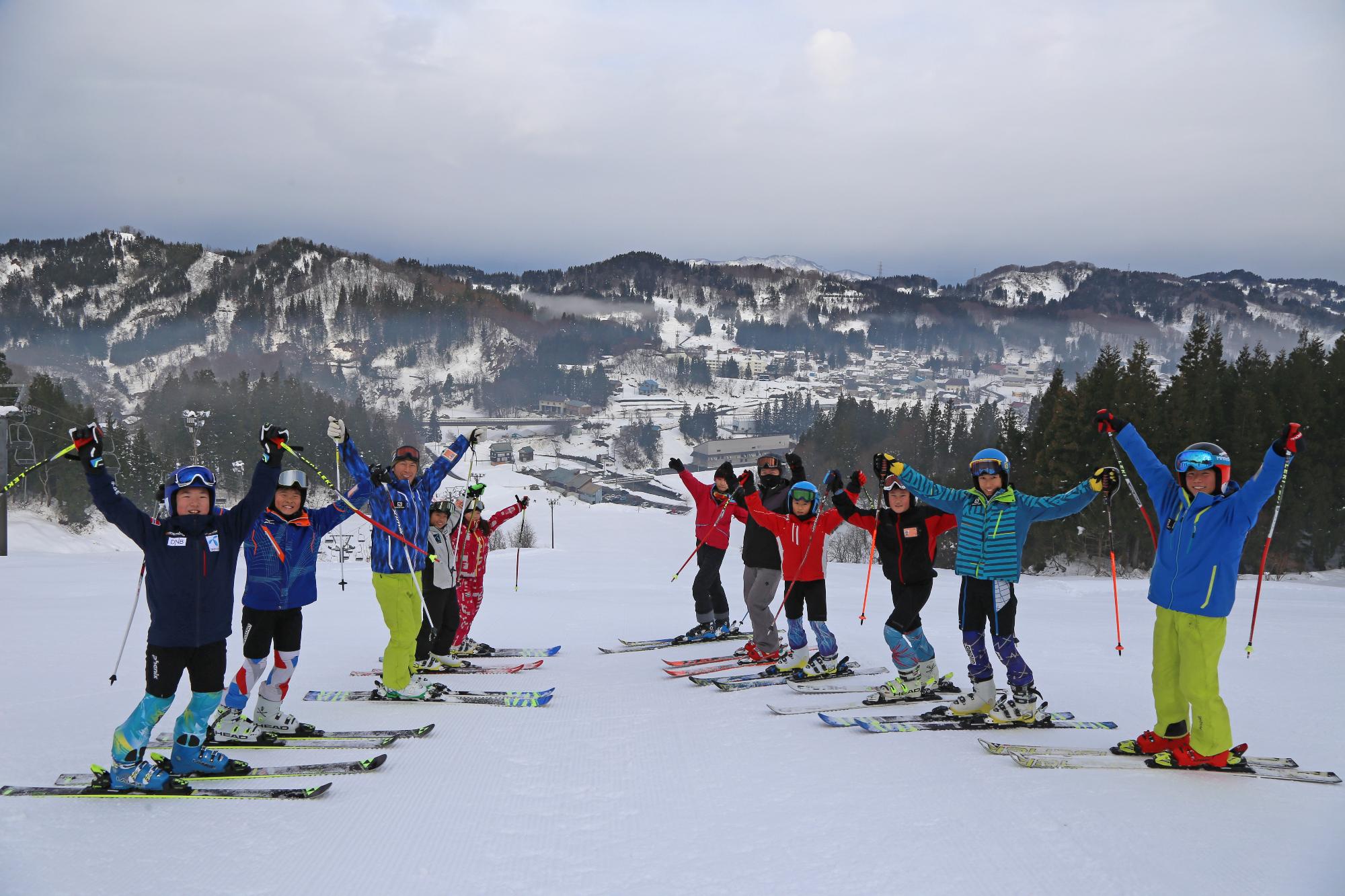 スキー場でスキー板を履いた状態で斜面を背に両手を上げてポーズをとる子どもたち
