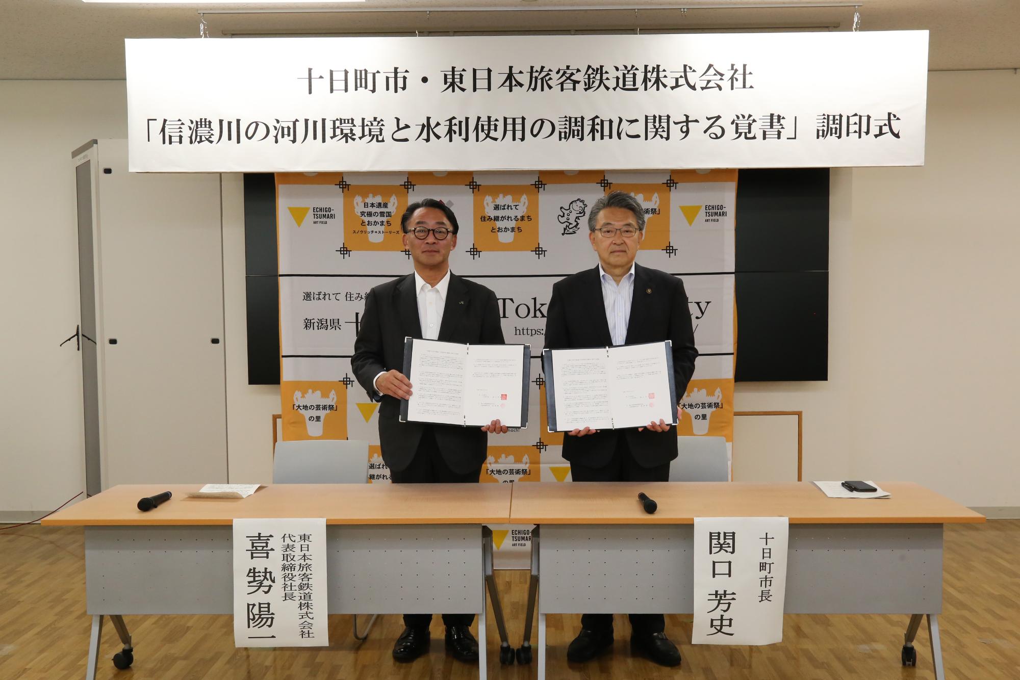 自治体のロゴが書かれた壁紙を背に覚書を見せる関口市長と喜勢社長の写真