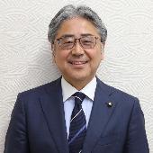 眼鏡をかけスーツを着て微笑んでいる富井高志議員の写真