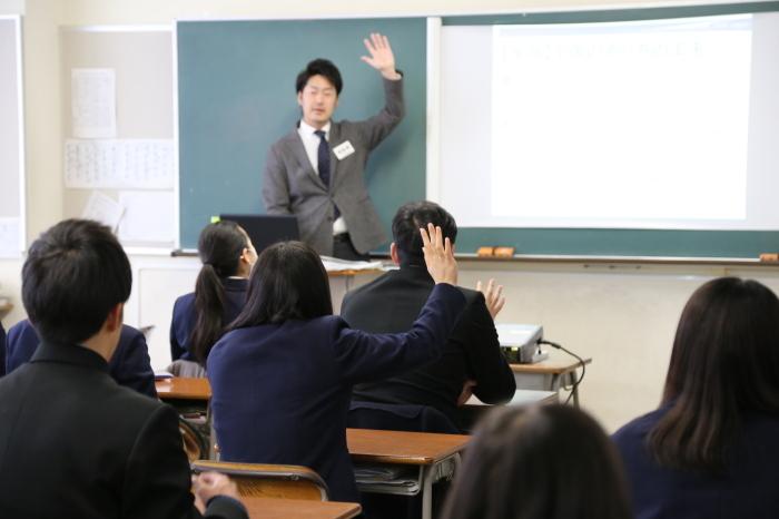 教室内で着席しておる学生の数人が手をあげている後ろ姿と黒板の前で立っているスーツの男性が左手をあげている写真