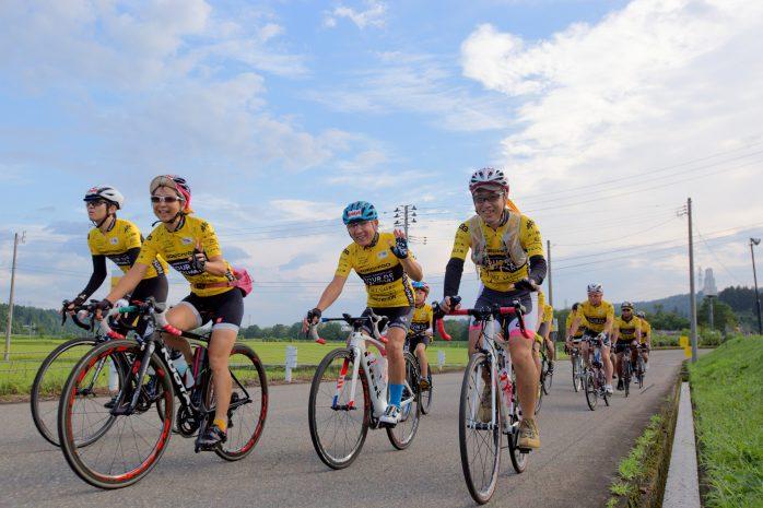 青空と水田を背景に黄色と黒の服装でサイクリングをしている人々の写真
