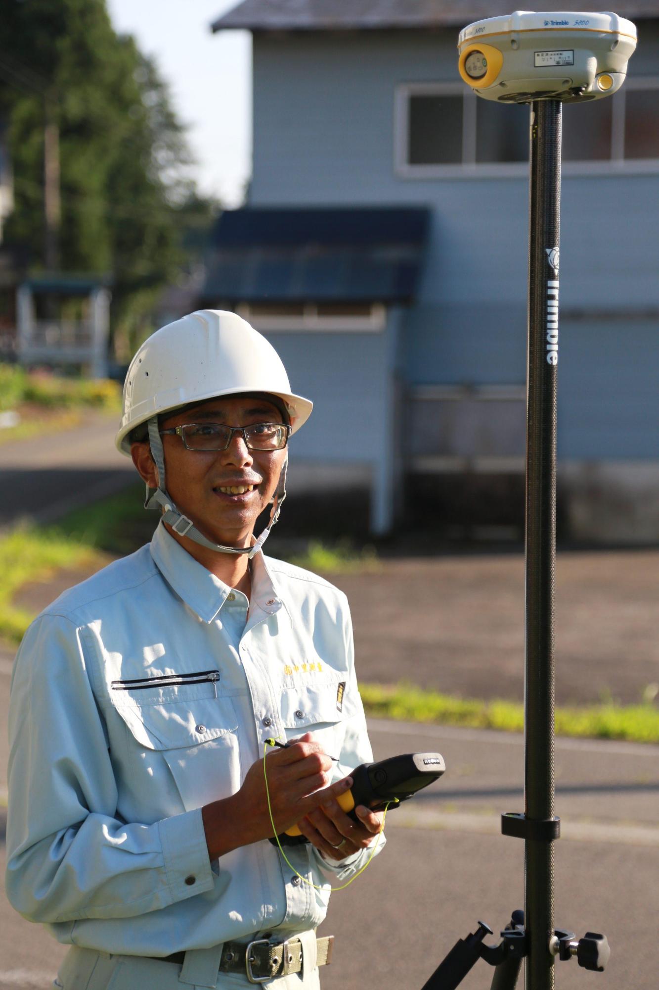 棒の先端に円形の機械が付いたGPS測量機の横で、作業着姿にヘルメットの眼鏡の男性がこちらに笑顔を向けている写真