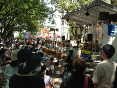 屋外ステージで太鼓のパフォーマンスを観覧する大勢の人々。