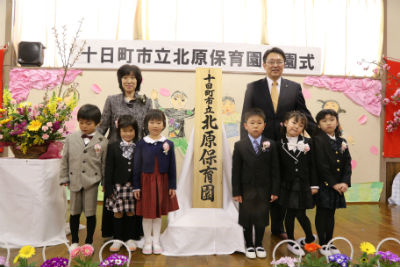保育園の看板の横で市長と子供たちが記念撮影している。
