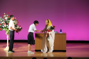 ステージ上で2人の生徒が美輪明宏さんに花束を贈呈している。
