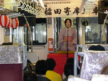 列車内の壇上で着物姿の女性が芸を披露している。