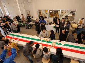 イタリア国旗のテーブルクロスが敷かれた長いテーブルを中心に周りで会食をする人々の写真