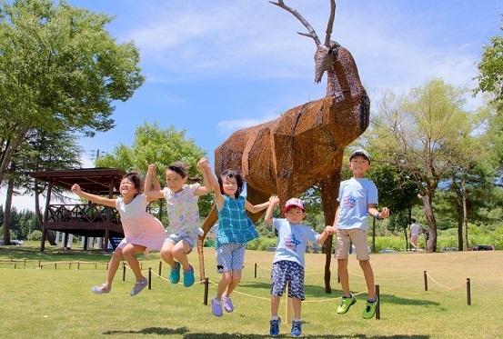鹿のような動物の彫刻の前で、子供たち5人が手をつなぎながらジャンプをしている写真