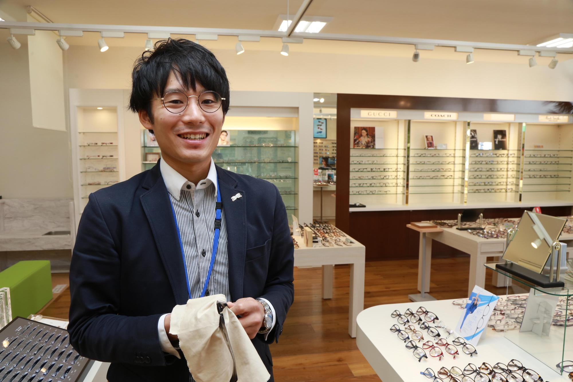 眼鏡が陳列されている机とともに写る笑顔の高橋雅人さんの写真