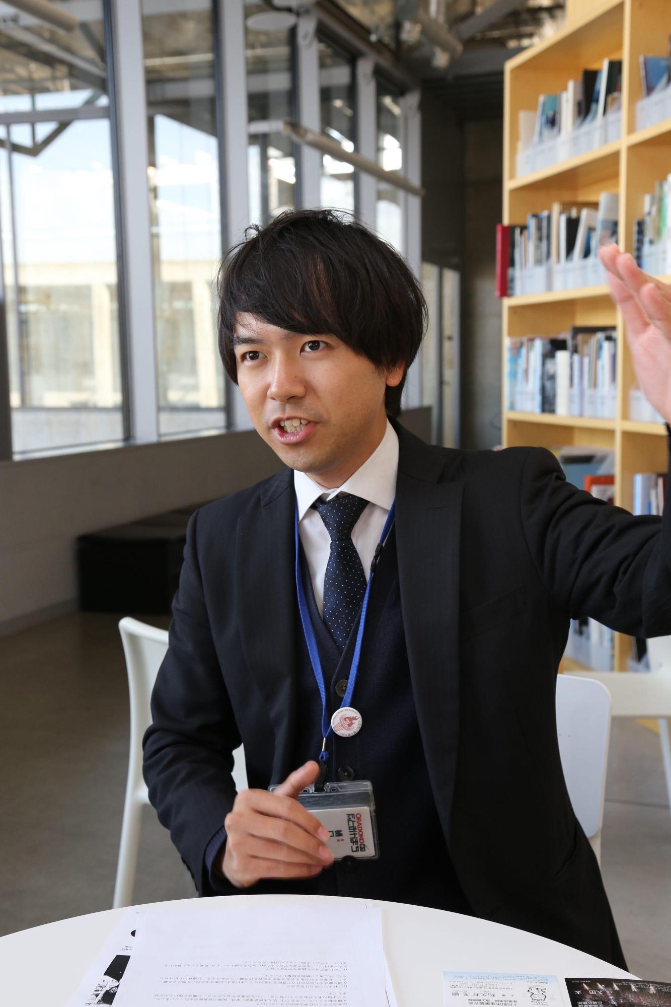 白い円形のテーブル席でスーツ姿の樋口智史さんが左手を挙げ業務内容を説明している写真