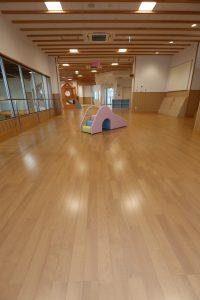 木目調の床の広い空間の中央に幼児向けの小型の滑り台が設置されている「はいはい広場」内観の写真
