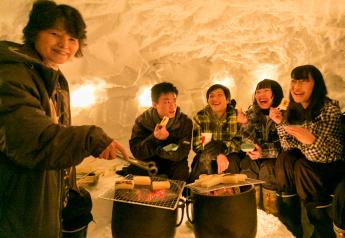 雪の家の中でもちを焼いて食べている様子の男女5人組の写真