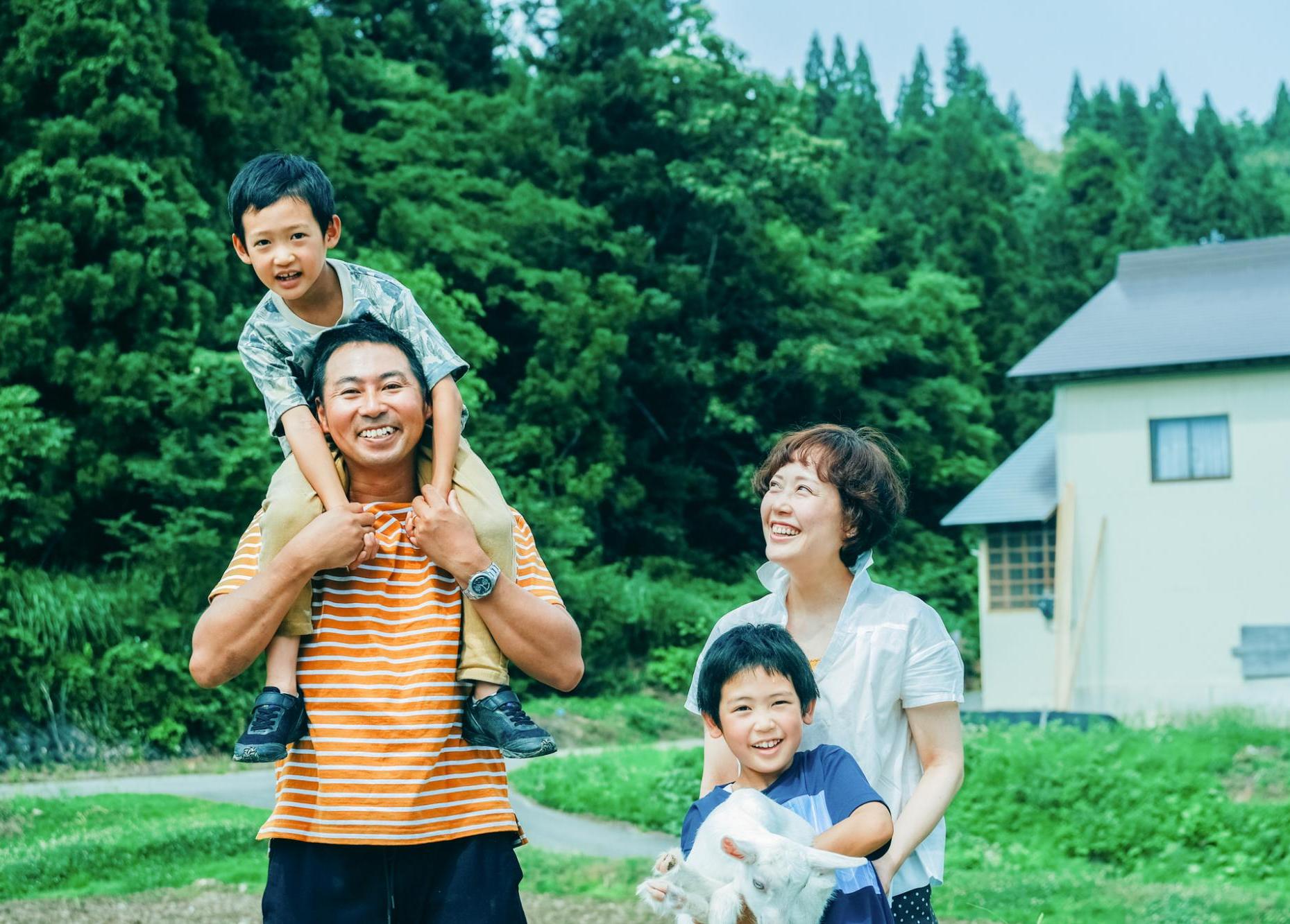 家族4人とヤギが森の前で笑顔でいる