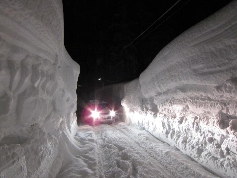 両サイドに雪が高く積んで車一台分の横スペースを通る道を作り、その道を通る車の写真