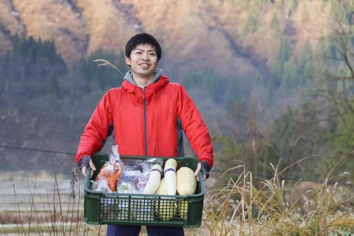 山あいの中で、コンテナに入った沢山の野菜を笑顔でこちらに見せている赤いジャンパーの男性の写真