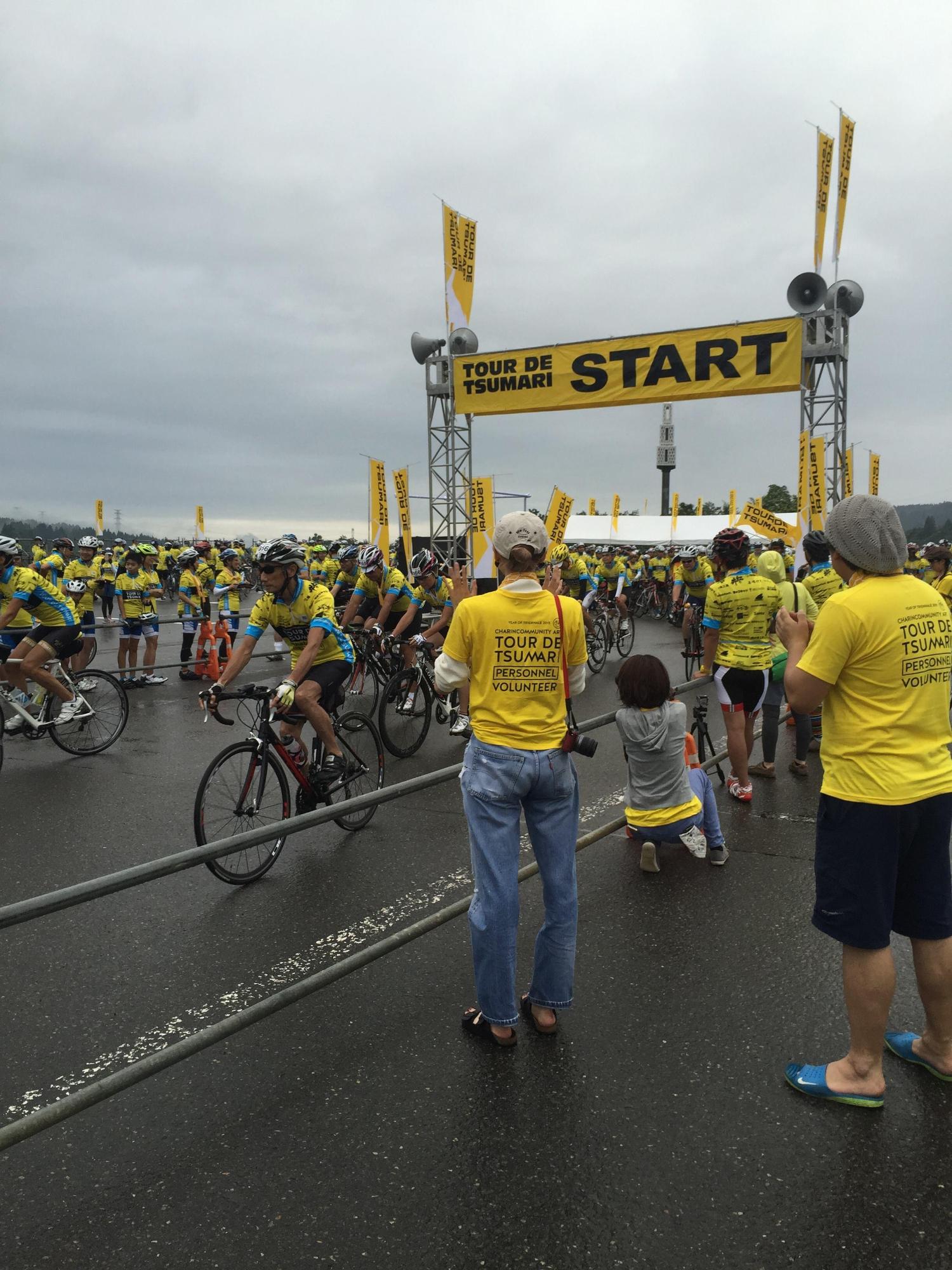 黄色い旗が掲げられたスタートのゲートから、自転車競技の参加者達が一斉に走り出してくる様子の写真