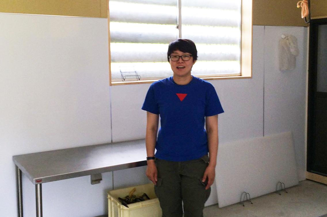 白い壁にシルバーの台が置かれている室内で、青いTシャツを着て立ってこちらに笑顔を向ける、ショートカットの高橋美佐子さんの写真