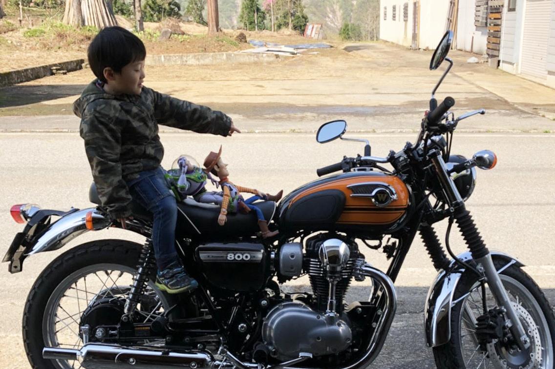 道路の上で、黒い車体にオレンジの差し色の入った大型2輪バイクにぬいぐるみと一緒にまたがっている、迷彩服の小さな男の子の写真