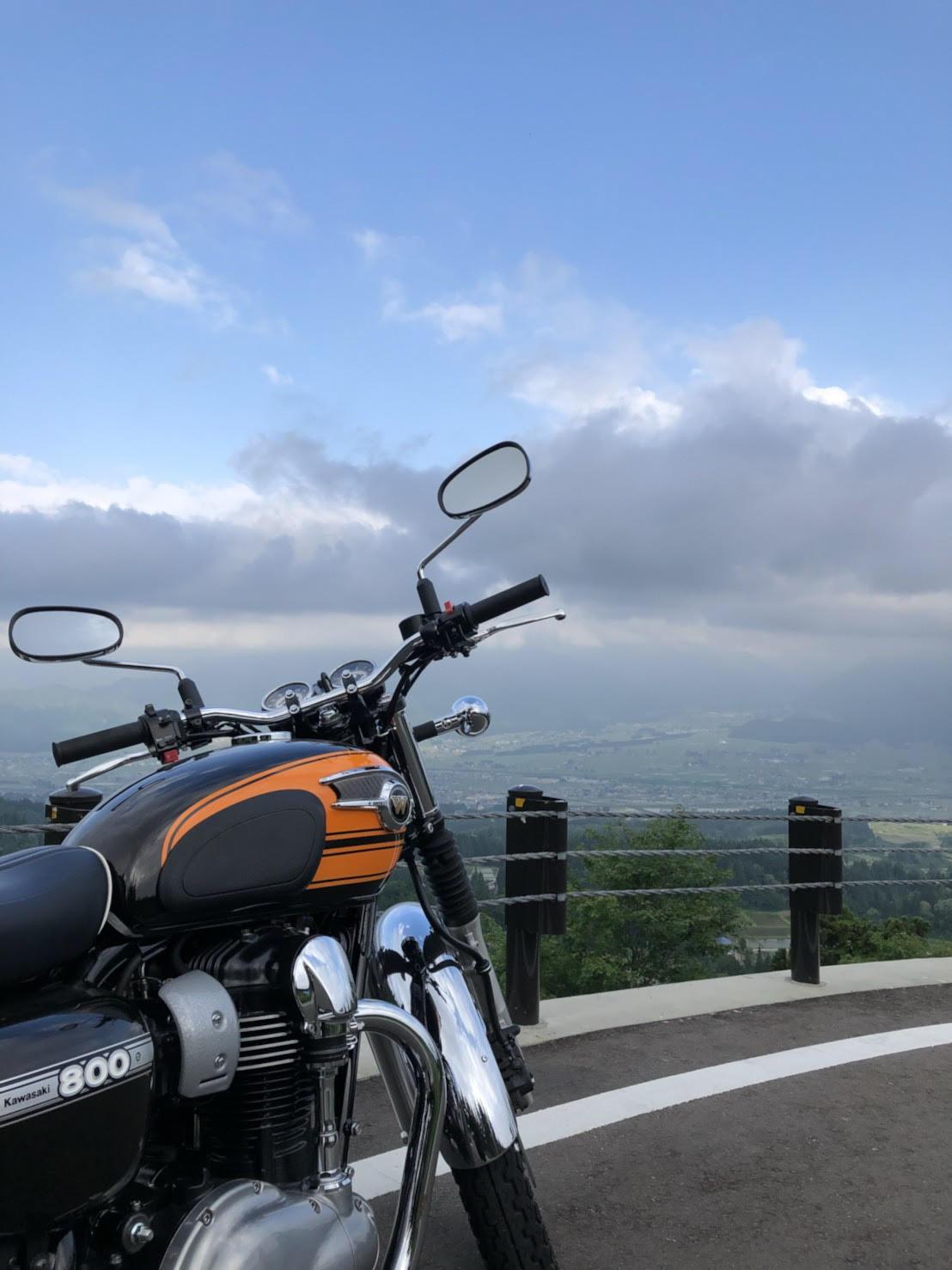 緩やかなカーブを描く車道に止められた大型2輪バイク。バイク越しに見える、高台からの緑の多い町の風景と、大きな雲が浮かぶ青空の写真