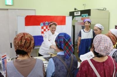 エプロンと三角巾をつけた参加者たちが先生から料理の説明を受けている写真