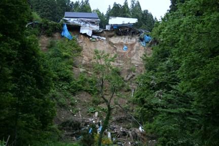 上に崩壊した建物が2つあり、下が崖のようになっていて土がむき出しになっている、樽沢集落の崩落箇所の写真