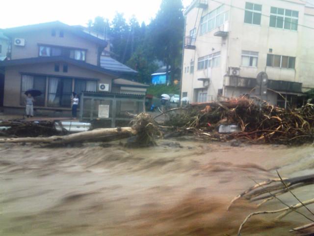 氾濫した河川が木々を押し流して民家の前を激しく流れているところを住人が心配そうに見ている写真